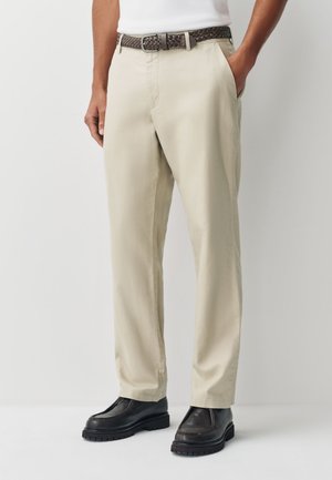 Mann trägt beige gerade geschnittene Hose mit Seitentaschen, einen braunen gewebten Gürtel und schwarze Lederloafers, steht vor einem einfachen Hintergrund.