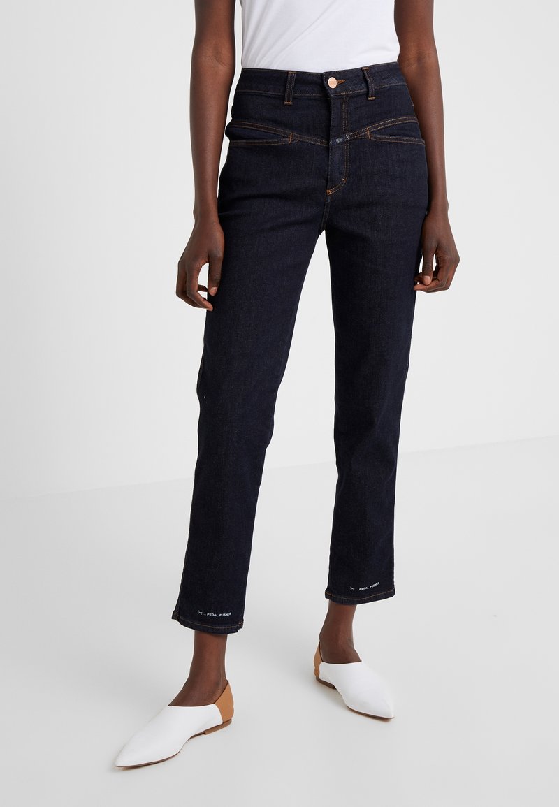 Personne portant un jean droit taille haute bleu foncé et des ballerines blanches pointues à talons beiges debout sur un sol clair.