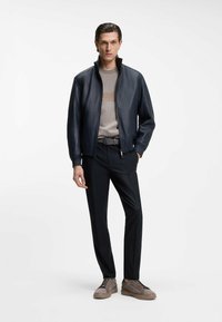 Μπλε δερμάτινο μπουφάν με φερμουάρ, γκρι πουλόβερ με στρογγυλή λαιμόκοψη, μαύρο παντελόνι και καφέ αθλητικά παπούτσια με υφασματικές λεπτομέρειες και κορδόνια.