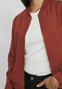 Rostfarbener Bomberjacke mit goldenen Reißverschluss, geripptem Kragen und Bündchen. Getragen über einem weißen gerippten Oberteil, kombiniert mit schwarzen Hosen.