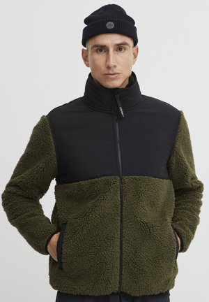 Fleecejacke mit einem schwarzen oberen Abschnitt und einem tiefgrünen unteren, mit einem Reißverschluss und Seitentaschen. Die Textur ist weich und flauschig.