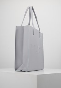 Jasnoszara torba tote Ted Baker London z podwójnymi uchwytami, stojąca pionowo na białym piedestale na jednolitym tle.
