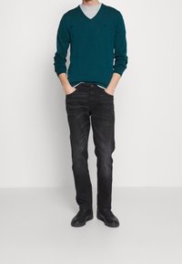 Mężczyzna stoi z rękami w kieszeniach, ubrany w turkusowy sweter w serek, białą koszulkę T-shirt pod spodem, czarne dżinsy i czarne sneakersy na jednolitym tle.