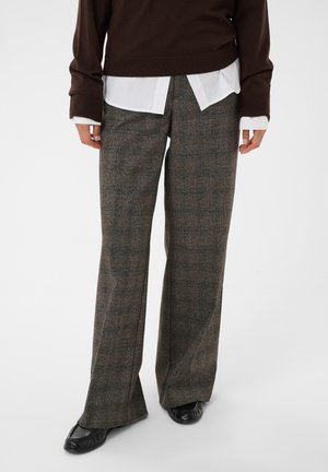Personne portant un pull marron, une chemise blanche, un pantalon large en tweed et des mocassins noirs, debout sur un fond uni.