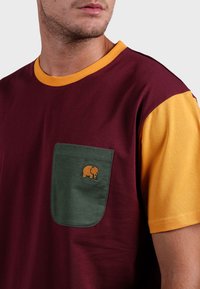 Chemise à manches courtes avec un corps bordeaux, des manches jaunes et une poche verte. La poche est ornée d'une petite broderie d'éléphant orange.
