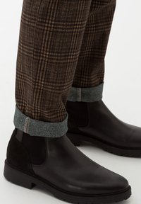 Pantalon à carreaux marron retroussé au-dessus de bottines Chelsea en cuir noir avec talon en daim et panneaux latéraux élastiques sur fond blanc.