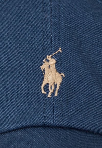 Broderie beige d’un joueur de polo à cheval tenant un maillet levé sur un fond de tissu bleu, probablement une casquette.