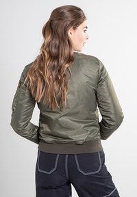 Olijfgroene bomberjack met een glanzende, doorgestikte afwerking, geribbelde manchetten en zoom, en een zijvakdetail op de mouw. Draagt donkere broek.