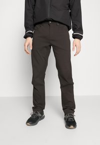 Tamno smeđe hlače uskog kroja, izrađene od rastezljivog materijala. Imaju četiri džepa i standardni pojas s petljama za remen.