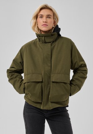 Mujer rubia con chaqueta acolchada verde oliva con cuello alto y grandes bolsillos delanteros, de pie con las manos en los bolsillos frente a un fondo gris.