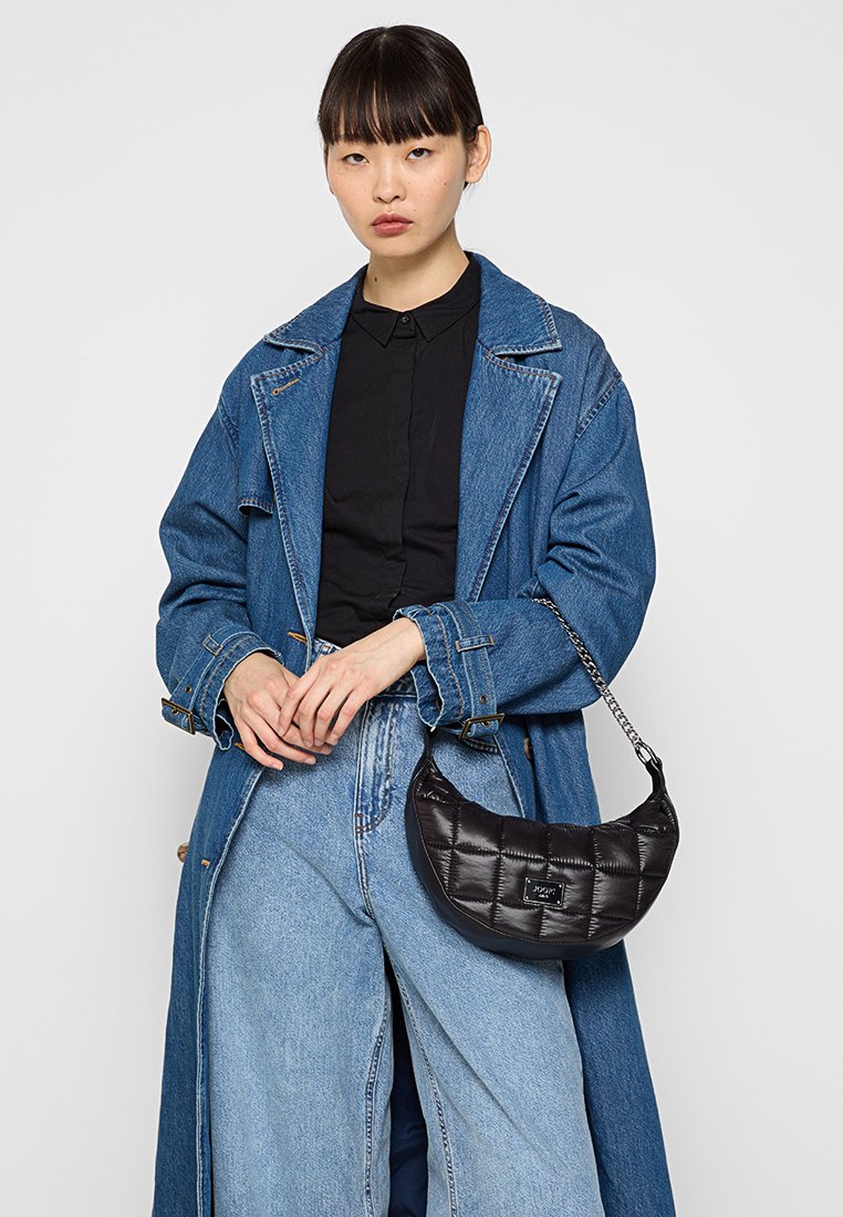 Schwarze gesteppte Handtasche mit geschwungener Form, ausgestattet mit einem kleinen Markenschild und einer Kettenakzentuierung. Tragen mit einem langen Denim-Mantel und weitem Jeans.