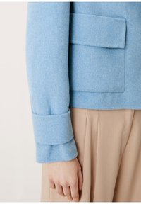 Ljusblå ullblazer med vikta ärmar och ficka, tillsammans med beige plisserade byxor som visar en jämn struktur.