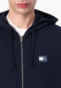 Marineblå zip-hoodie med foret hætte, der har en frontlomme og et "TOMMY JEANS" logo-patch i blå og hvid på brystet.