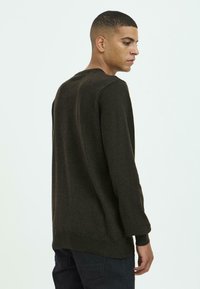 Jonge man met kort haar, gekleed in een donkere lange mouwen trui en donkere broek, kijkt opzij tegen een effen lichte achtergrond.