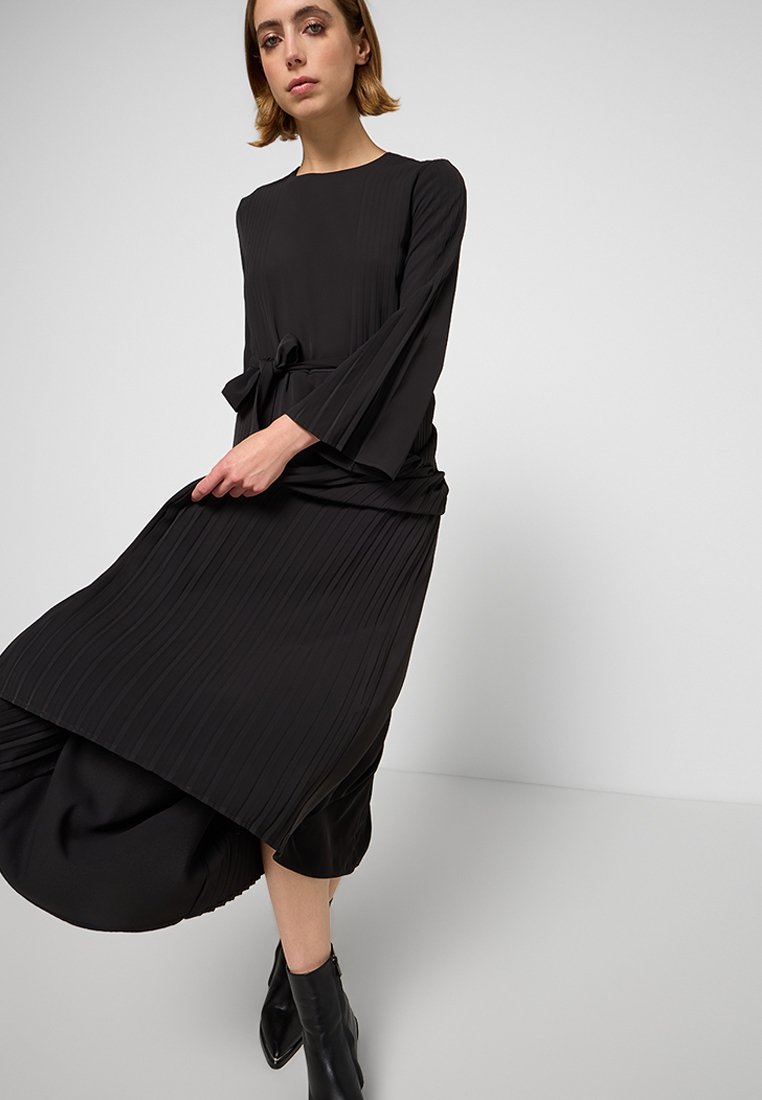 Abito nero plissettato con maniche lunghe, scollo rotondo e cintura in vita. Tessuto liscio con orlo a più strati e dettagli di design minimi.