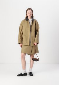 Olivgrön jacka med svart krage, matchad med en kjol i samma färg. Modellen håller en liten brun väska. Texturerat tyg, avslappnad passform.