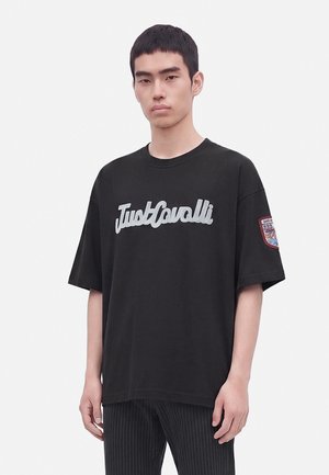 Млад мъж, облечен с черна овърсайз тениска с думите "Just Cavalli" отпред и нашивка на ляв ръкав.