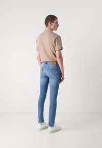 Camisa polo beige, jeans ajustados de color azul claro y zapatillas blancas. Las características notables incluyen una etiqueta de cuero marrón en la cinturilla de los jeans.