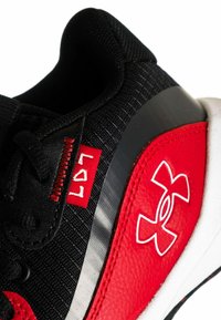 Zapatilla deportiva negra con acentos rojos, parte superior texturizada, franja plateada y logo de Under Armour. Diseño de caña alta, construcción de material resistente.