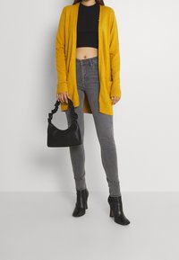 Gul lång kofta, svart crop top, grå skinny jeans, svarta klackboots och en svart handväska med strukturerat handtag.