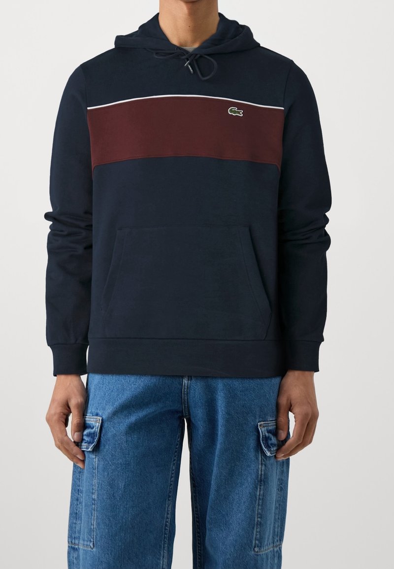 Homme portant un sweat à capuche bleu marine avec une bande bordeaux sur la poitrine et un petit logo crocodile vert, associé à un jean cargo bleu.