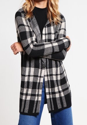 Femme portant un cardigan à carreaux noir et blanc boutonné sur un haut noir et un jean bleu, les bras croisés, sur fond blanc.