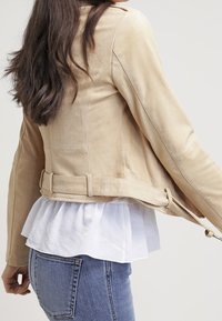 Donna che indossa una giacca scamosciata beige sopra una blusa bianca con volant e jeans blu, mostrata di spalle e di lato con i capelli sciolti.