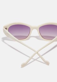 Valged cat-eye päikeseprillid Gradient-lilla läätsedega. Omavad õhukesi, siledad raame ja kaardus templeid, mis ristuvad taga.