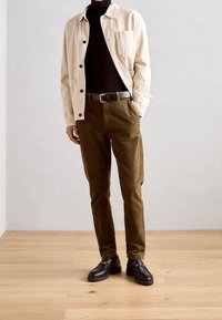 Beige button-up jacket over a black turtleneck, paired with olive green corduroy pants and black loafers with decorative hardware.