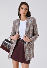 Geruite blazer in beige en bruin, gecombineerd met een witte t-shirt en een bordeauxrode plissérok. Bevat een handtas met een gestructureerd krokodillenpatroon.