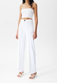 Biały dopasowany crop top zestawiony z białymi spodniami o wysokim stanie i szerokich nogawkach, które mają z przodu guzik. Przezroczyste sandały na obcasie na bosych stopach.