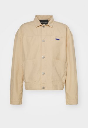 Beige cotton jacket with a collared neckline, silver snap buttons, two front pockets, and a small blue label on the left chest.