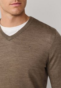 Bruine V-hals trui gemaakt van een gestructureerd materiaal, met een subtiele geborduurde embleem op de linkerborst. Draagt over een wit shirt.