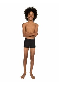 Shorts de bain noirs avec une ceinture logo, torse nu et cheveux bouclés. Debout, bras croisés et souriant sur un fond blanc.