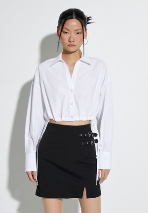 Camicia bianca cropped con bottoni, maniche lunghe e orlo con coulisse, abbinata a una gonna nera con spacco laterale e dettagli con fibbia.