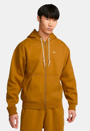 Brun zip-up hoodie lavet af blødt stof, med kangaroo-lomme og hvide snore. Inkluderer et lille Nike-logo på brystet.