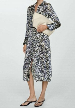 Kvinde iført en langærmet, mønstret midi-kjole med frontslids, holder en stor beige clutch og har sorte hæle på.