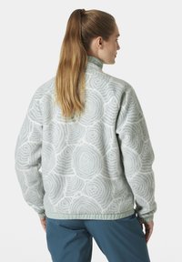 Kurtka polarowa w jasnoszarym kolorze z okrągłym wzorem drewna. Posiada wysoki kołnierz i elastyczny dół, prezentując miękki, teksturowany materiał.