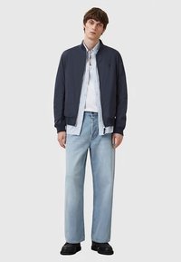 Noor mees, seljas must bomber-jakk helesinise nööpidega särk, valge T-särk, helesinised teksad ja mustad kingad, seisab püsti.