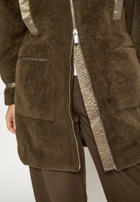 Hnedý flísový kabát so saténovými detailmi, ktorý má zapínanie na zips vpredu, dve predné vrecká a textúrovanú vnútornú podšívku.