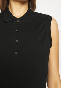 Ärmlös svart polo med fyra knappar och en liten broderad krokodil-logga på vänster bröst, som bärs av en person med ljus hy.