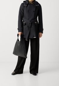 Manteau noir à double boutonnage, avec accents de boutons et ceinture. Pantalons larges noirs et sac cabas en cuir noir. Chaussures à bout carré.