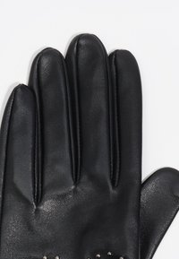 Guante de cuero negro con detalles visibles de costuras y pequeñas tachuelas de metal cerca de la muñeca sobre un fondo blanco.