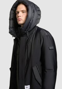 Chaqueta de plumón negra con una gran capucha acolchada. Cuenta con un cierre frontal con cremallera, bolsillos laterales y un parche del logotipo en la manga. Textura suave.