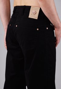 Schwarze Jeans aus Denim mit einer figurschmeichelnden Passform. Merkmale ein Lederpatch an der Taille, kupferfarbene Beschläge und zwei Gesäßtaschen mit Nahtdetails.