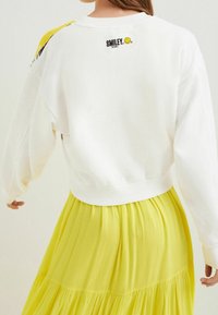 Bílá mikina s kulatým výstřihem a dlouhými rukávy, s žlutým detailem na rameni a černým logem "SMILEY" na zádech.