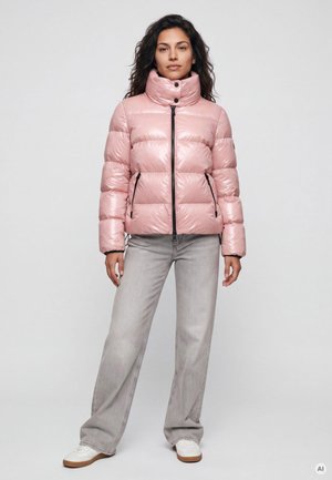 Frau trägt eine glänzende pinke Steppjacke, graue Jeans mit weitem Bein und weiße Sneakers, steht vor einem schlichten hellen Hintergrund.