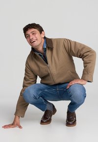 Młody mężczyzna klęczy na jednej ręce, ubrany w brązową kurtkę, dżinsową koszulę i dżinsy oraz brązowe skórzane buty, uśmiecha się i patrzy w lewo.