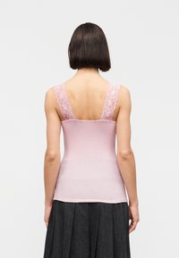 Γυναίκα με κοντά σκουρόχρωμα μαλλιά που φοράει αμάνικη ανοιχτό ροζ μπλούζα με δαντελένιους ιμάντες και σκοτεινή πλισέ φούστα, εμφανιζόμενη από πίσω.