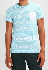 Camiseta de manga corta azul claro con un patrón geométrico blanco y un bolsillo en el pecho de color blanco sólido. Fabricada en tela suave.